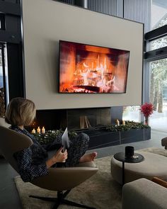 a woman sitting in a chair reading a book while looking at the fire on the fireplace