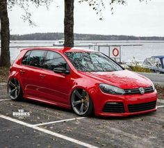 a red car parked in a parking lot next to some trees and boats on the water