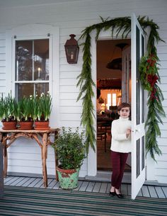 a woman standing in front of a white house with potted plants on the porch