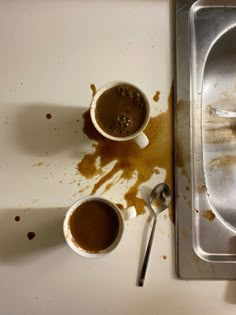 two cups of hot chocolate sit on a dirty counter next to a metal tray with spoons