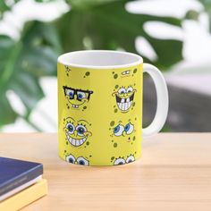 a yellow mug with cartoon faces on it sitting on a table next to a book