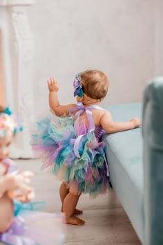 a baby girl in a tutu and dress standing on a blue couch next to another child