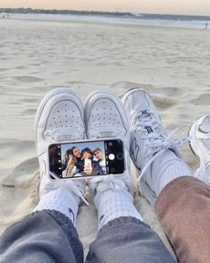 someone taking a selfie with their cell phone on the beach while wearing white sneakers