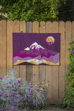 a purple quilt hanging on the side of a wooden fence with flowers in front of it