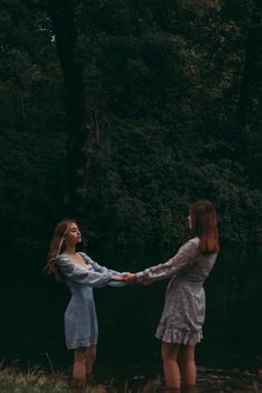 two young women holding hands in front of a body of water with trees behind them