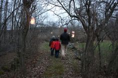 two people walking down a path in the woods with lanterns hanging from trees on either side