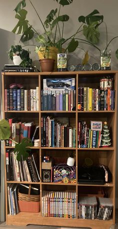 Light brown bookshelf. The bookshelf is one of those grided ones where each shelf is a square. 4 squares tall, 3 squares wide, 12 squares in total. fairly neatly organized. Lots of books and video games on their own compartments of the shelf. One shelf has a cute basket. A couple shelves have some plants, there is also larger plants ontop of the book shelf, aswell as some decorative coins? It looks very pretty overall. Mildly cottagecore. Open Shelf Bookshelf, Bookshelf Maximalist, Open Bookcase Styling, Wooden Bookshelf Aesthetic, Diy Vintage Bookshelf, Small Apartment Library, Trinket Bookshelf, 70s Bookshelf, Brown Bookshelf Decor