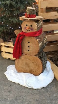 a snowman made out of logs sitting in front of a christmas tree with a red scarf around its neck