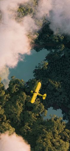 an aerial view of a yellow plane flying over trees and water in the sky with clouds