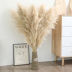 a vase filled with dry grass next to a white wall and wooden floored area