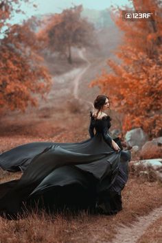 a woman in a long black dress is walking through an autumn forest with her hair blowing in the wind