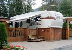 a white trailer parked in front of a wooden fence with flowers and trees around it