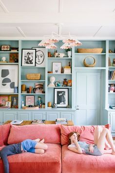 two people laying on a pink couch in front of bookshelves with pictures above them