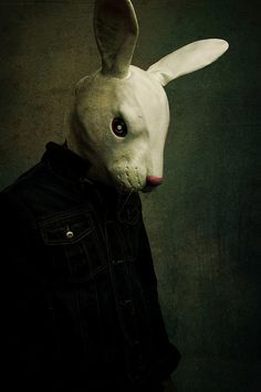 a white rabbit wearing a black jacket and standing in front of a dark background with the head of an animal on it's face