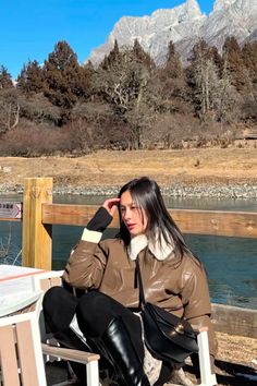 a woman sitting on top of a wooden bench next to a body of water with mountains in the background