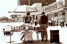 two people standing in front of a table with electronics on it and an umbrella over them