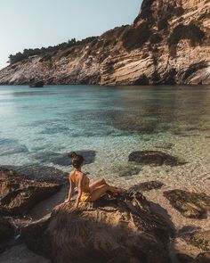 a woman sitting on top of a rock next to the ocean with clear blue water