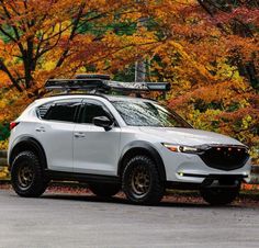 a white mazda car parked in front of trees with fall leaves on the ground behind it