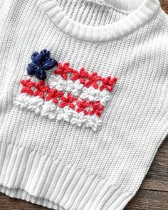 a white sweater with red, white and blue crochet on the front is laying on a wooden surface