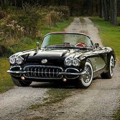 an old black car parked on the side of a dirt road next to a forest