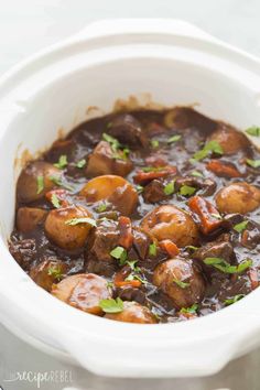 a white crock pot filled with beef and potatoes covered in gravy, garnished with parsley