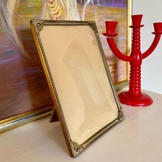 a red candle holder next to a painting on a table with a framed picture in front of it