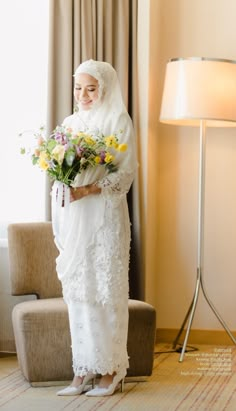 a woman in a hijab holding flowers standing next to a chair and lamp