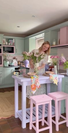 a woman standing at a kitchen island with two stools in front of it and flowers on the table