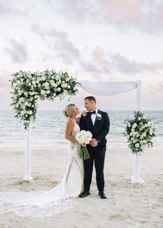 a bride and groom standing on the beach under an arch decorated with white flowers for their wedding ceremony