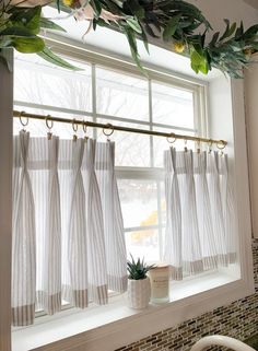 a kitchen window with white curtains and green plants hanging from the windowsill, next to a potted plant