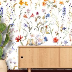 a wooden cabinet sitting in front of a wall with flowers on it and a plant next to it