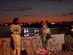 two people standing next to each other near a wall with graffiti on it and the sky in the background