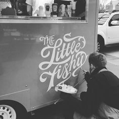 black and white photograph of a man painting the side of a food truck with lettering on it