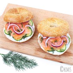 two bagels with salmon, cucumber and onions on them sitting on a cutting board