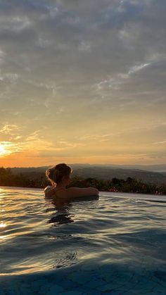 a woman sitting in the middle of a swimming pool at sunset with her back turned to the camera