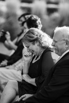 an older couple laughing while sitting on a bench in front of some other people and taking pictures with their cell phones