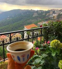 someone holding up a cup of coffee in front of a balcony overlooking the city below