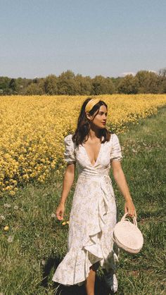 a woman in a white dress is holding a hat and walking through a field with yellow flowers