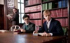 two young men sitting at a table in front of bookshelves, one holding a cell phone