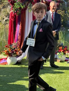 a young boy in a suit and tie walking on grass with other people behind him