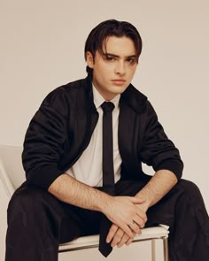 a man in a black suit and tie sitting on a chair with his legs crossed