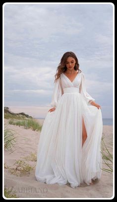 a woman standing on top of a sandy beach wearing a white dress and long sleeves