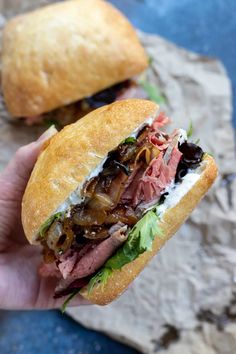 a hand holding a sandwich with meat, cheese and lettuce on it next to another sandwich