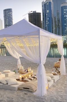 a white canopy bed sitting on top of a sandy beach next to tall city buildings