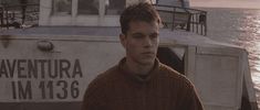 a young man standing in front of a boat on the water at sunset or sunrise