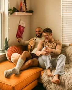 two men sitting on a couch with their dog and stockings hanging from the windowsill