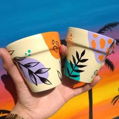 two hand painted cups are held up in front of a palm tree and beach scene