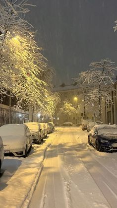 snow covered cars parked on the side of a snowy street at night with buildings in the background