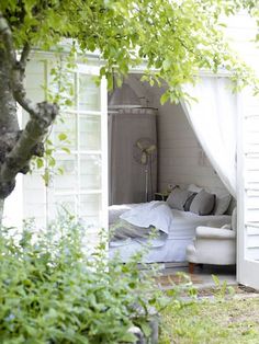 a bed sitting under a tree next to a white building with a curtain on it's side