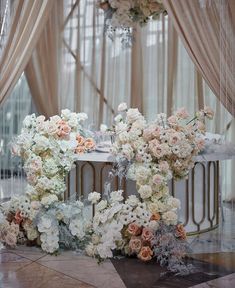 a table topped with lots of white and pink flowers next to a tall window covered in sheer drapes